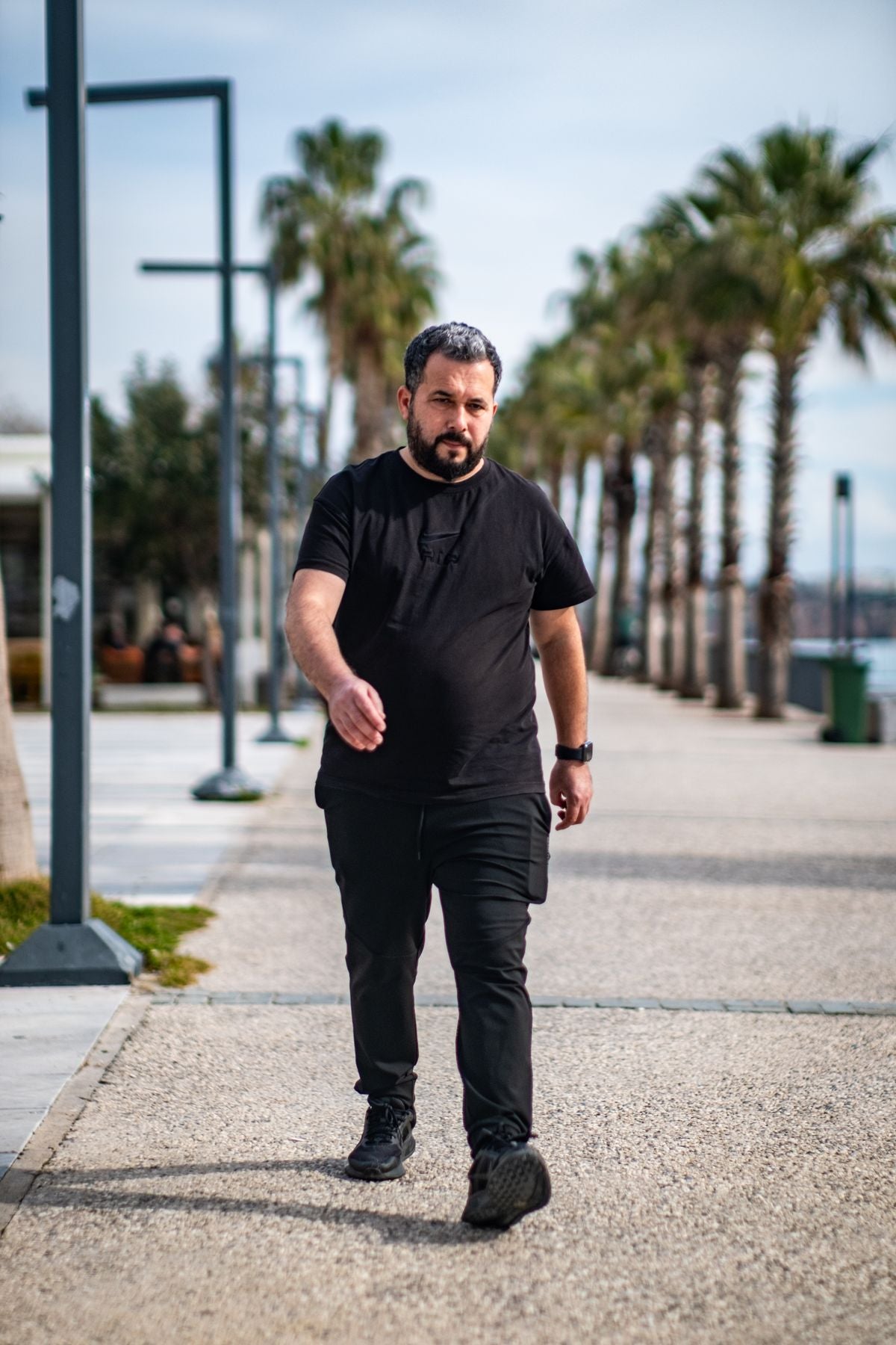 Stockier man in black crew neck tee with joggers walking on promenade — flattering fit on larger build